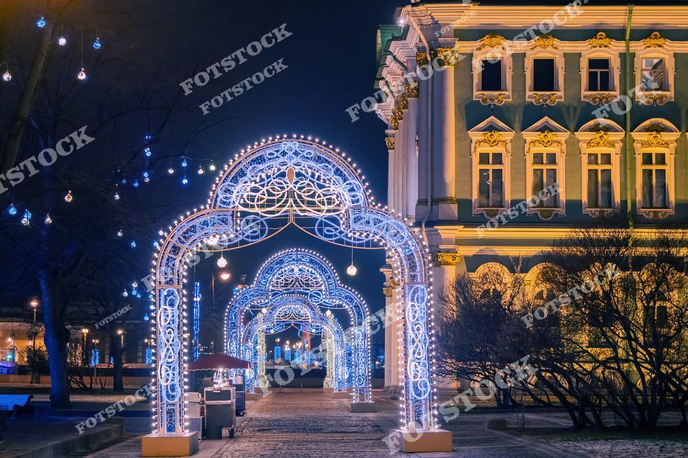 На фотографии изображена часть Дворцовой набережной рядом с Эрмитажем. Праздничные световые арки, украшенные гирляндами, создают атмосферу новогоднего волшебства. Архитектура Эрмитажа с подсветкой гармонично сочетается с праздничным убранством. Это место оживает в зимний вечер, привлекая жителей и гостей города.
